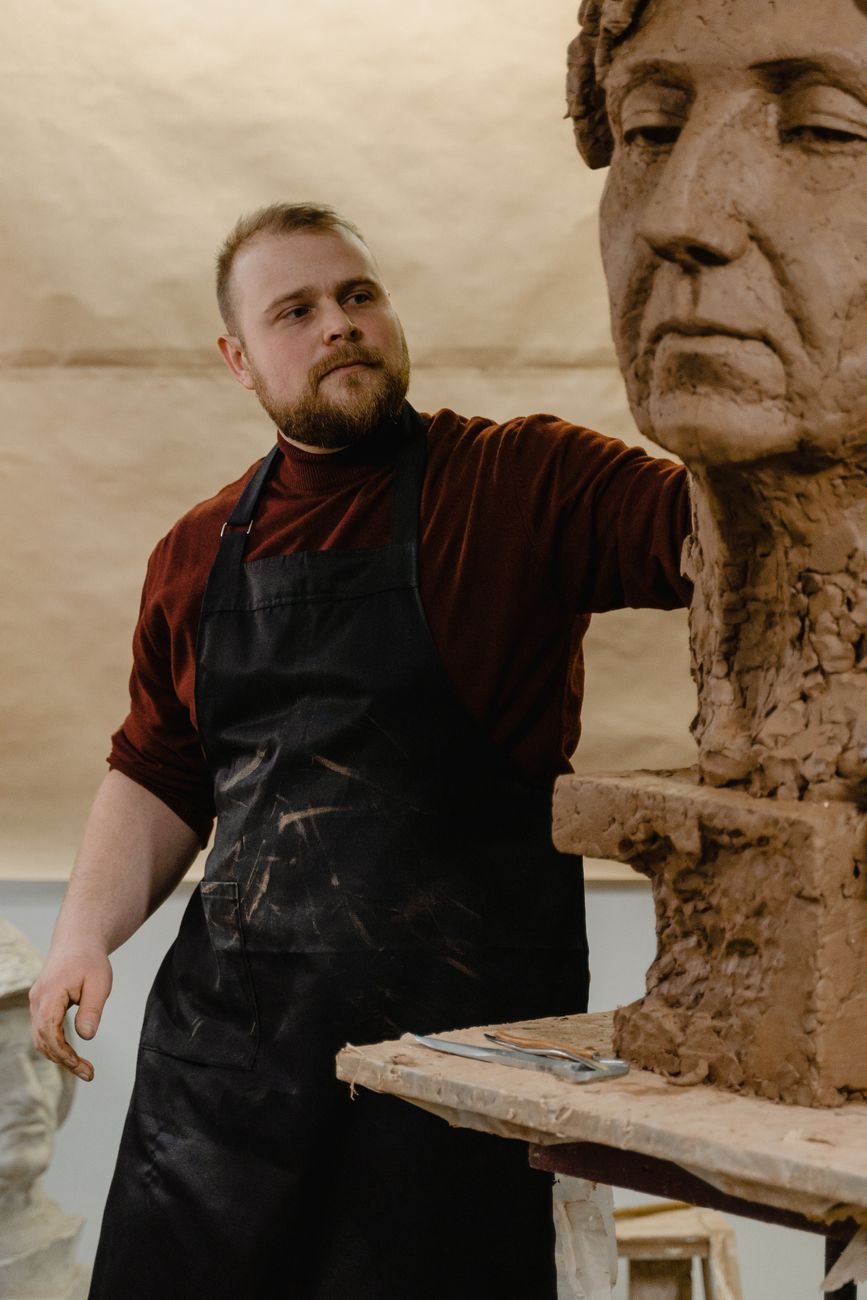Bearded male sculptor working on a large clay bust in art studio setting.